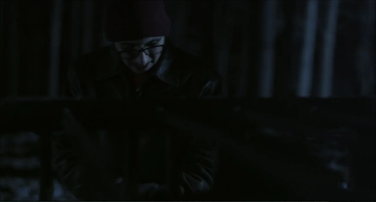 Freedom (2024) movie still: medium — A dark, minimally lit interior space features a person in shadows. The subject wears a beanie and glasses while…
