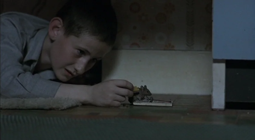 Ratcatcher (1999) movie still: closeup — A dimly lit domestic interior features patterned wallpaper and worn flooring. A young boy lies on his…
