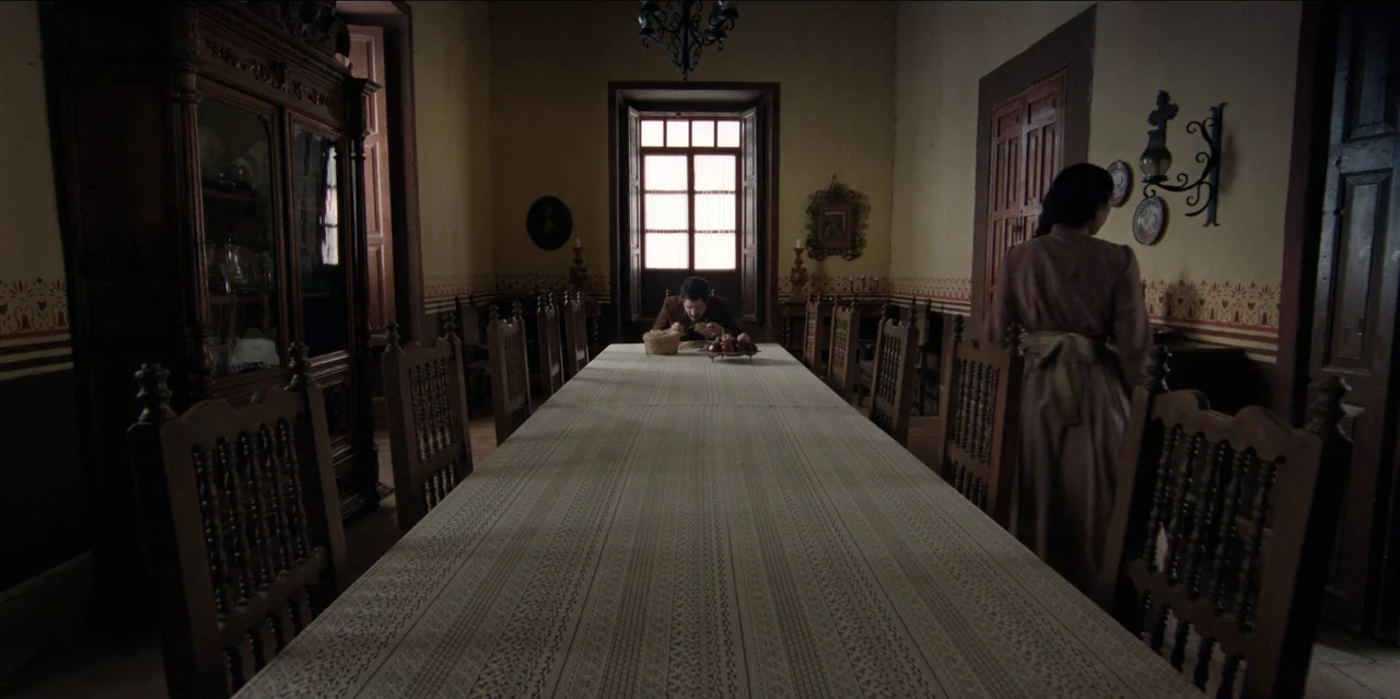 Pedro Páramo (2024) movie still: wide — A long dining room features dark carved wooden furniture and a large central window. A man eats at the far…