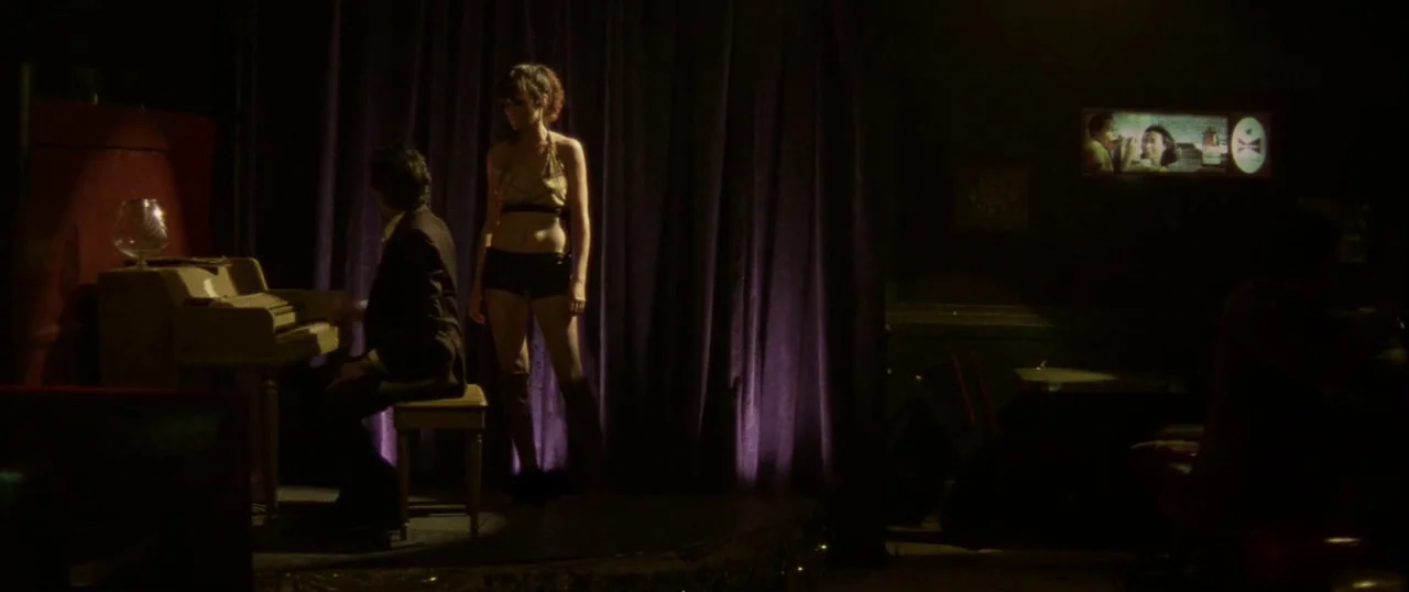 Low Down (2014) movie still: wide — A dimly lit lounge with deep purple curtains serving as a backdrop to a stage. A man in a suit plays a…