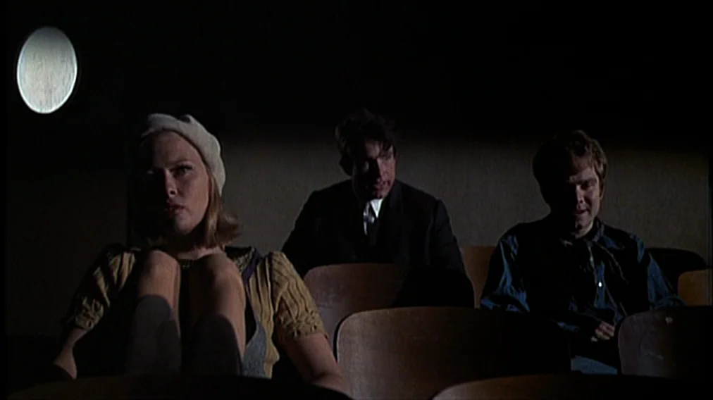 Bonnie And Clyde (1967) movie still: medium — Three people sit in a dark, empty cinema hall with rows of wooden chairs. The woman in the foreground…