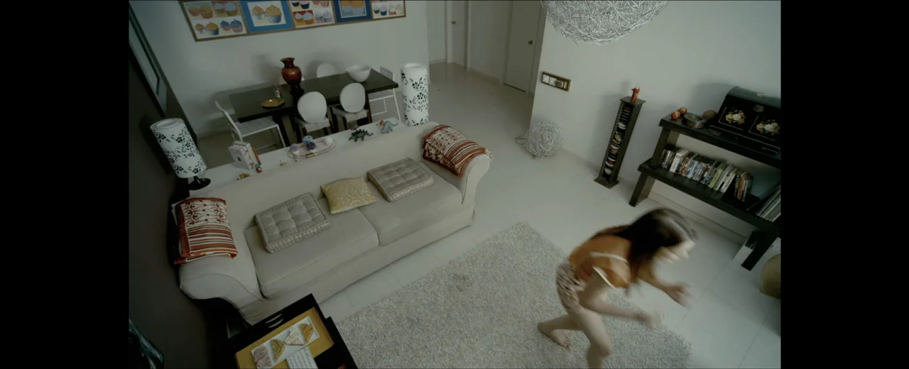 Faraday (2013) movie still: wide — A modern apartment with a beige couch and dining set in the background. A woman in a yellow shirt walks across a…