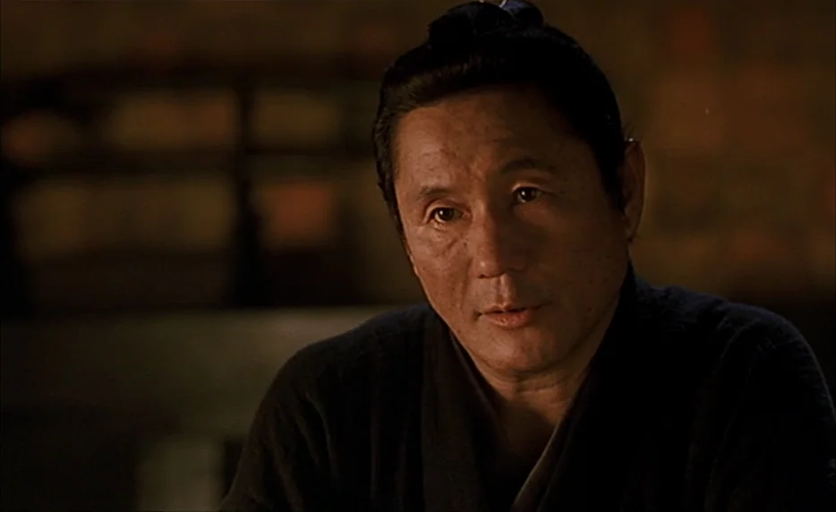 Gohatto (1999) movie still: closeup — A dimly lit room provides a dark, out-of-focus background for a portrait. An East Asian man in a traditional…
