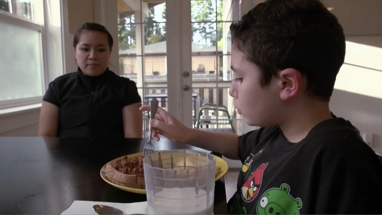 A Dangerous Son (2018) movie still: medium — A brightly lit indoor dining area features a dark table near glass sliding doors. A young boy eats a…