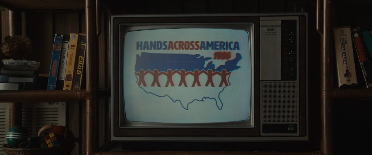Us (2019) movie still: closeup — A wooden entertainment center holds a vintage television showing a Hands Across America logo. Several VHS tapes are…