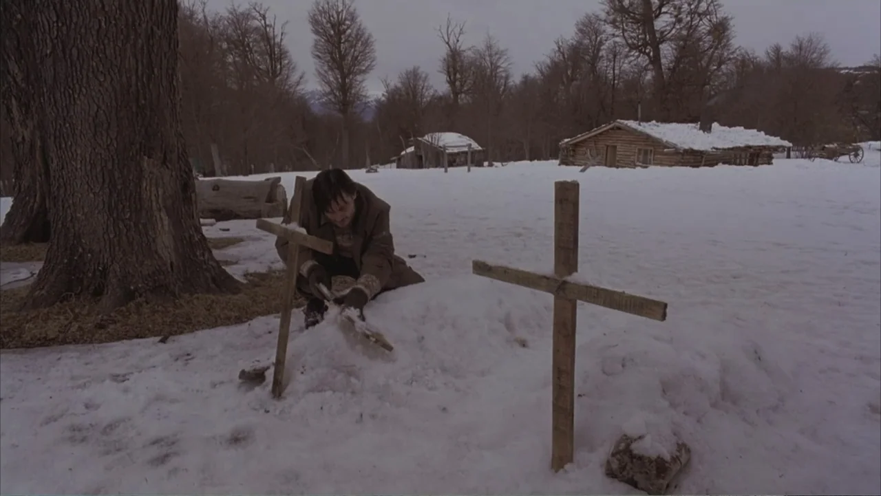 A Distant Place (2020) movie still: medium — A snow-covered clearing features several crude wooden grave markers and a distant log cabin. A man…