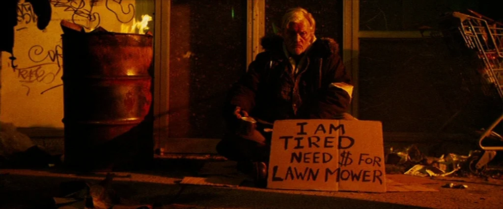 Hobo With A Shotgun (2011) movie still: medium — An older man sits on a concrete surface next to a burning metal barrel. He holds a hand-written…