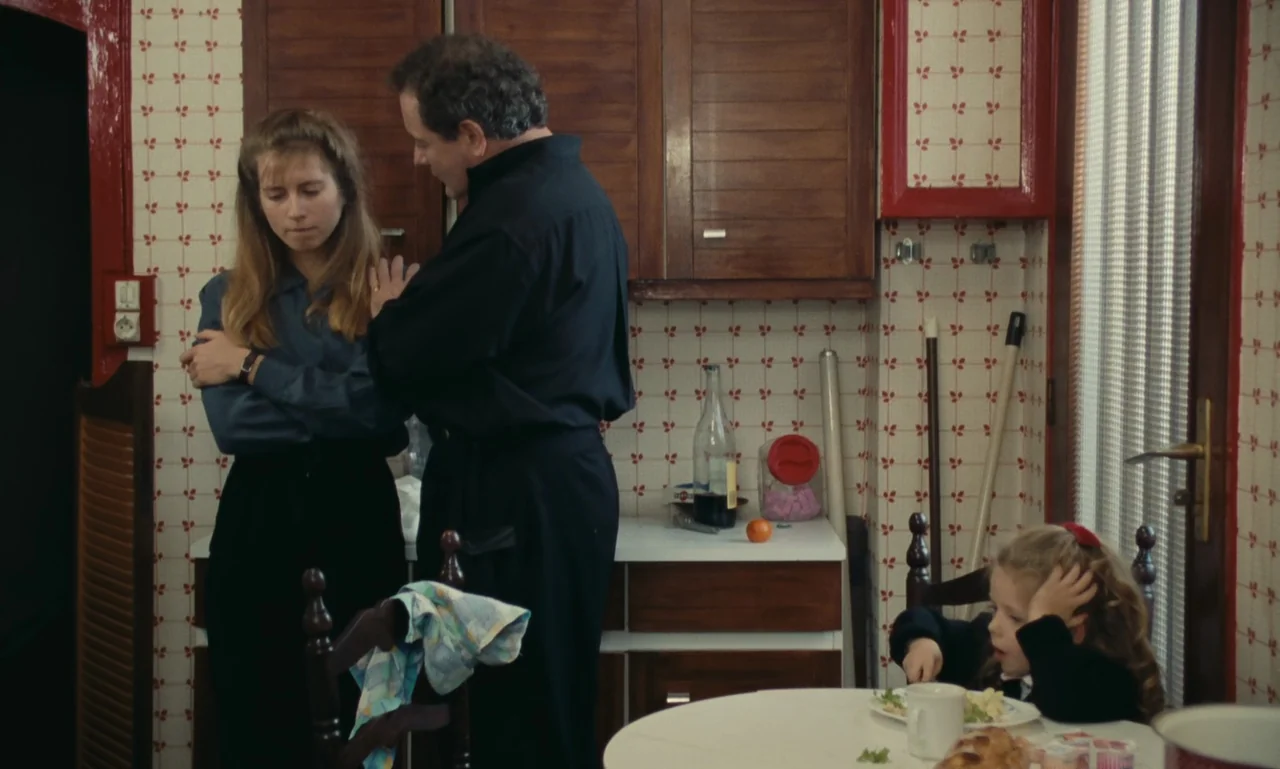A Tale of Winter (1992) movie still: medium — A dated home setting features patterned wallpaper and dark wooden cabinets above a counter. A man…