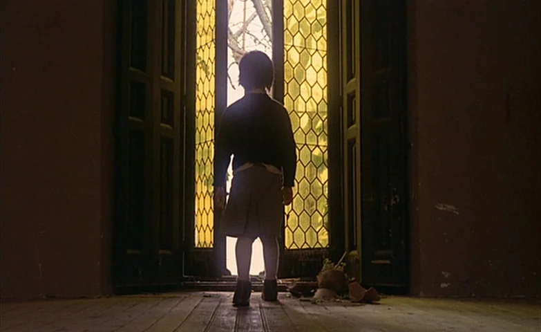 Spirit Of The Beehive (1973) movie still: fullbody — A dimly lit interior room features a child standing before a large open doorway. Sunlight…