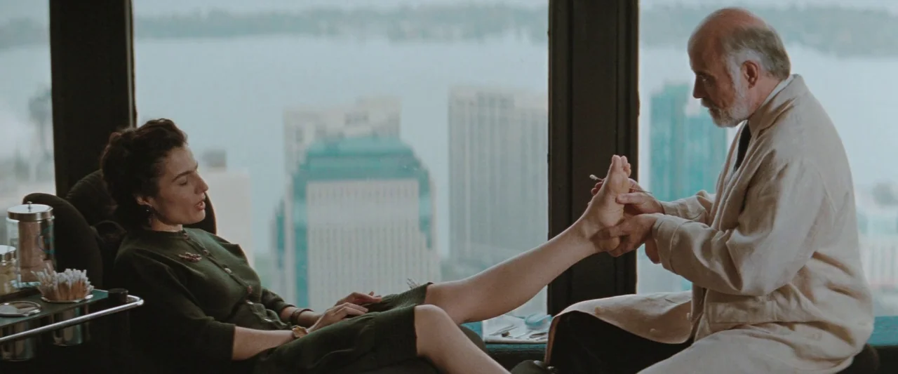 The Adjuster (1991) movie still: medium — A high-rise room with floor-to-ceiling windows overlooking a distant city skyline. A man performs a…