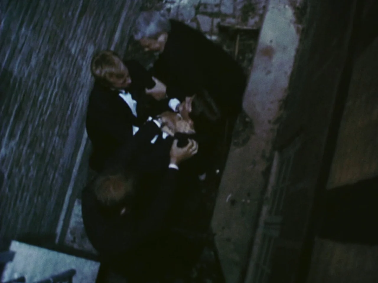 Festen (1998) movie still: medium — A narrow alleyway with stone walls at night is illuminated by overhead lighting. Three men in formal tuxedos…