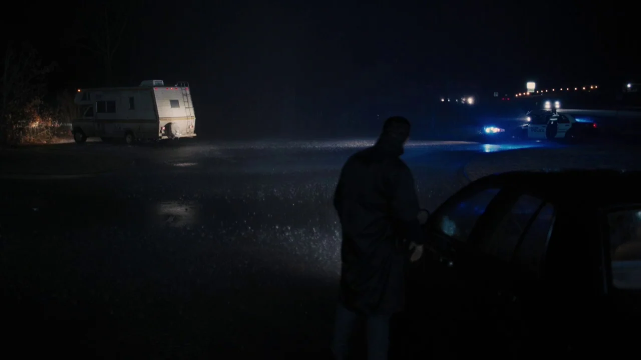 Prisoners (2013) movie still: wide — A rain-slicked dark parking lot at night features a distant recreational vehicle and police car. A man in a…