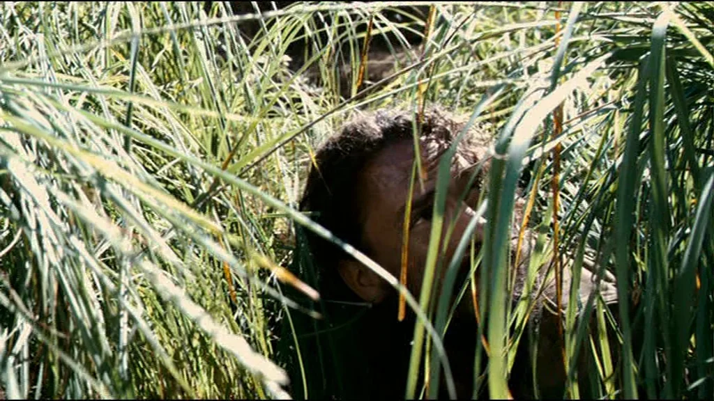 Che Part 2 (Guerrilla) (2008) movie still: closeup — A man hides within a patch of tall, dense blades of meadow grass. He peeks through the foliage…