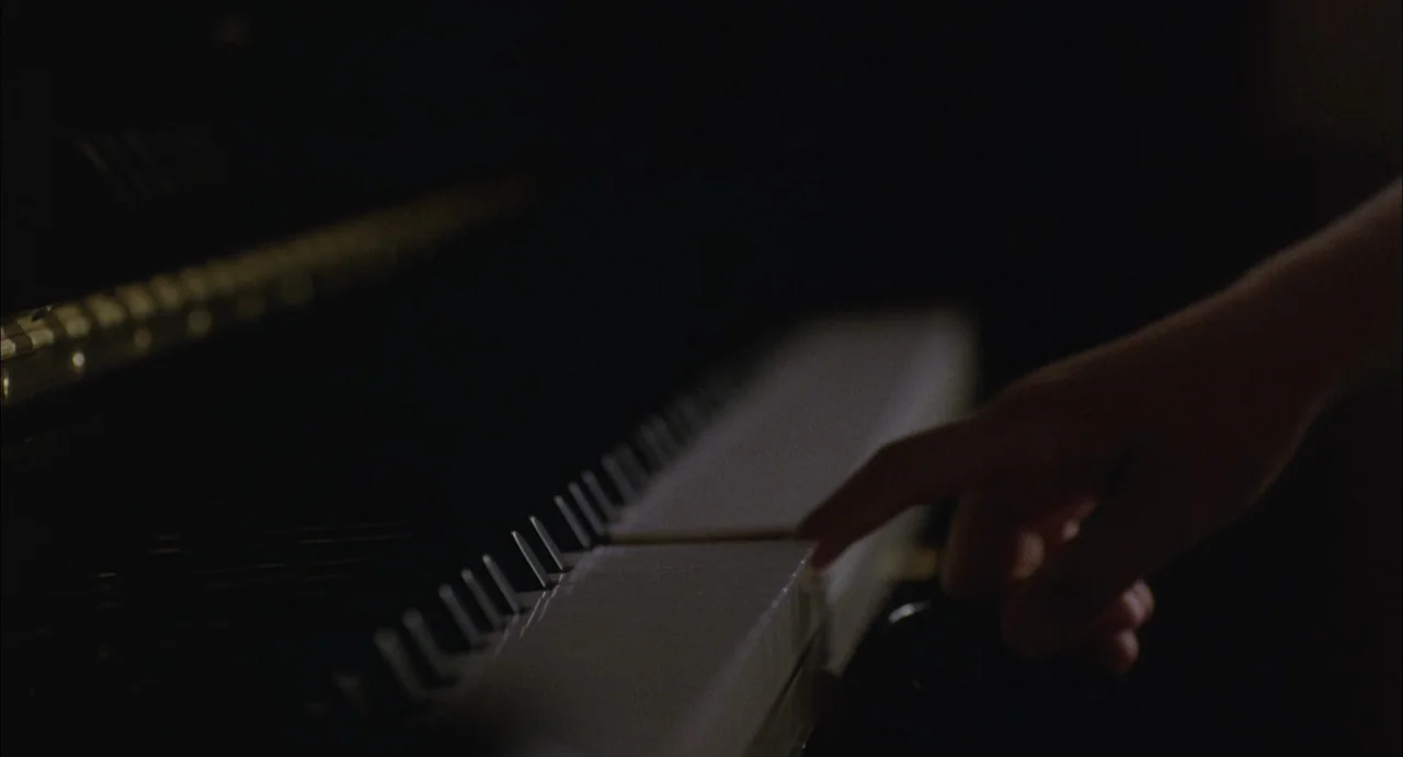 Still Walking (2008) movie still: closeup — A close-up view of a piano keyboard in a very dark room. A single hand touches a white piano key.