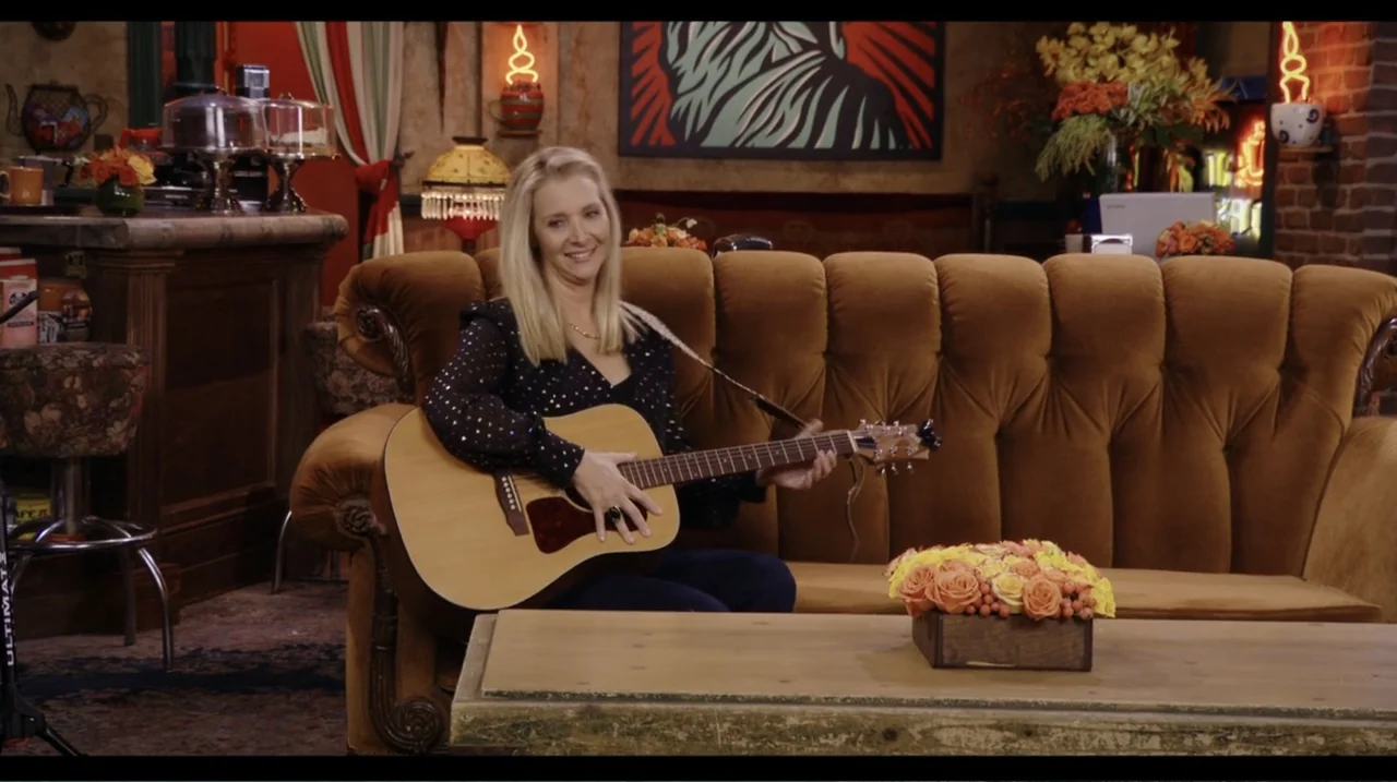 Friends: The Reunion (2021) movie still: medium — A 1990s coffee shop features an orange velvet sofa against a brick wall. A woman sits on the couch…