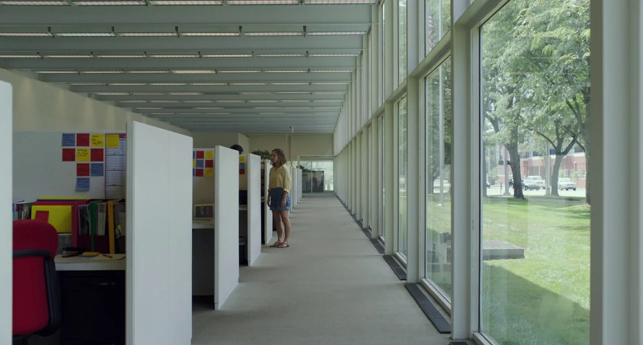Columbus (2017) movie still: wide — An open-plan workplace with white modular partitions and floor-to-ceiling windows overlooking a green lawn. A…