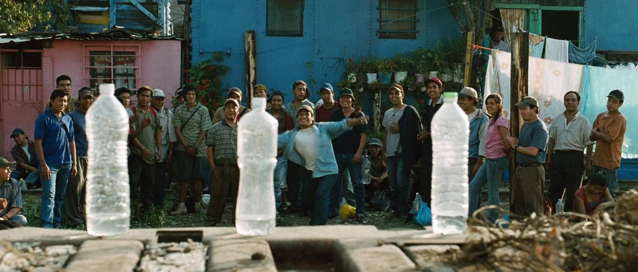 Sin Nombre (2009) movie still: fullbody — A group of people stands in front of blue and pink wooden houses. A young man throws an object toward…