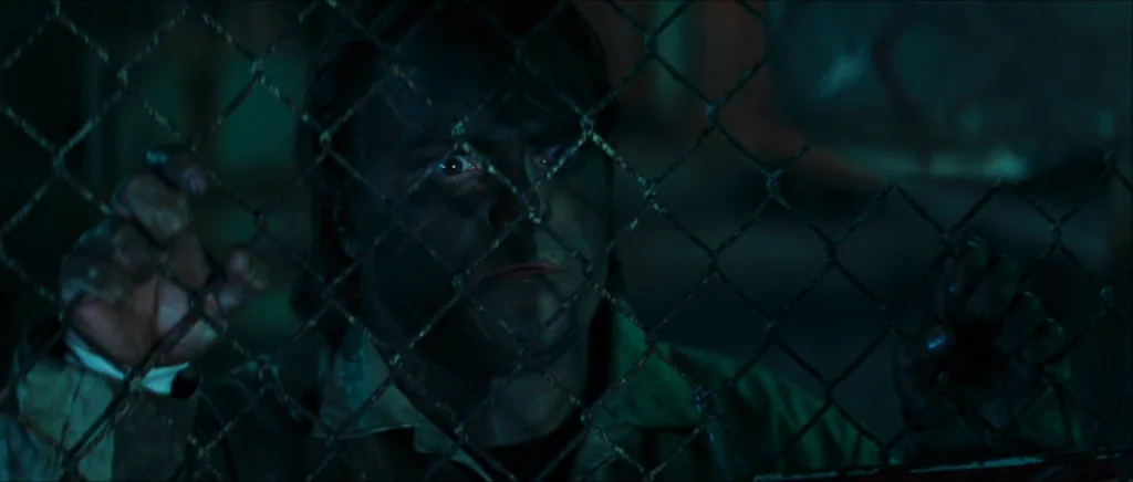 Be Kind Rewind (2008) movie still: medium — A man stands behind a metal mesh grid in darkness. He grips the wire barrier with both hands, looking…