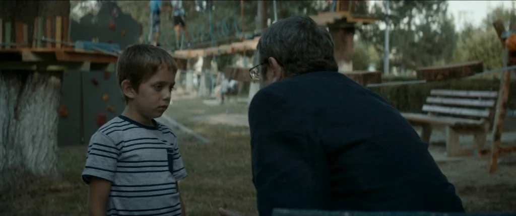 Graduation (2016) movie still: medium — An outdoor playground features wooden climbing structures and benches in a park setting. A small boy stands…