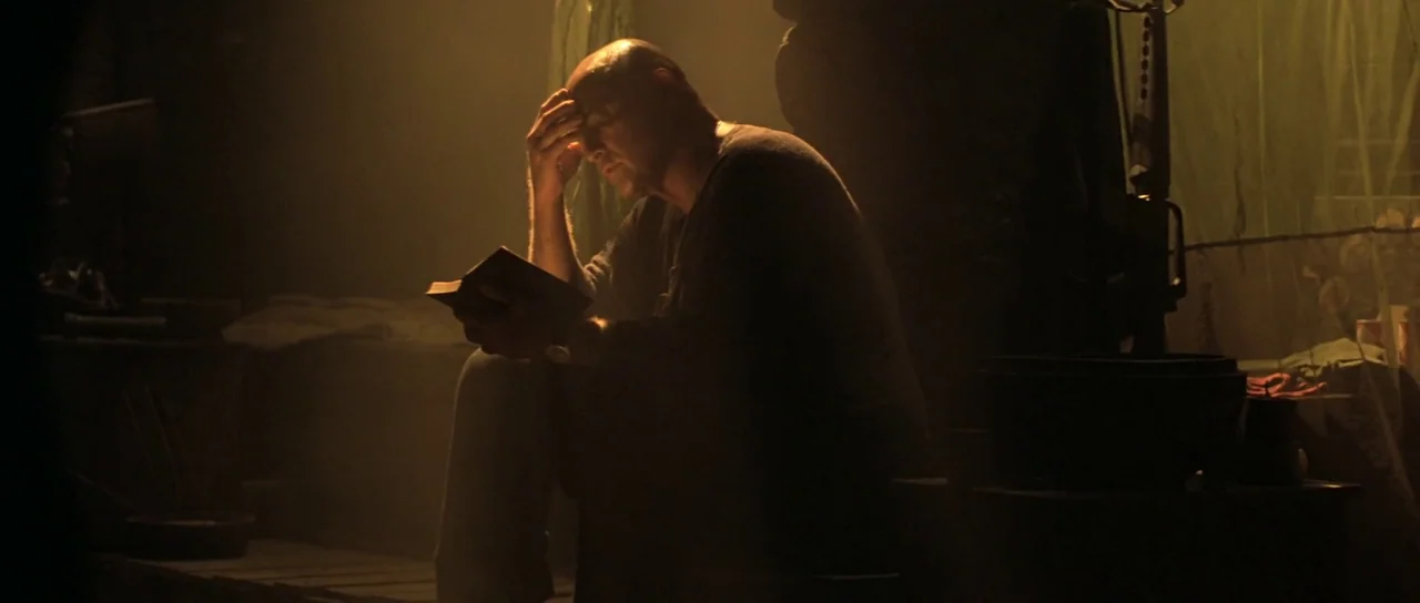 Apocalypse Now (1979) movie still: medium — A dark, dimly lit military space features a man seated in a wooden area. The man reads a small book…