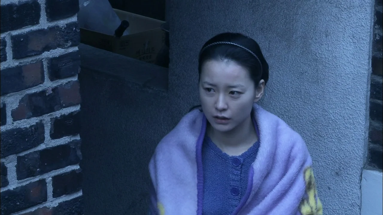 Oki’s Movie (2010) movie still: medium — A woman sits against a gray concrete corner next to a brick wall. She is wrapped in a lavender blanket…