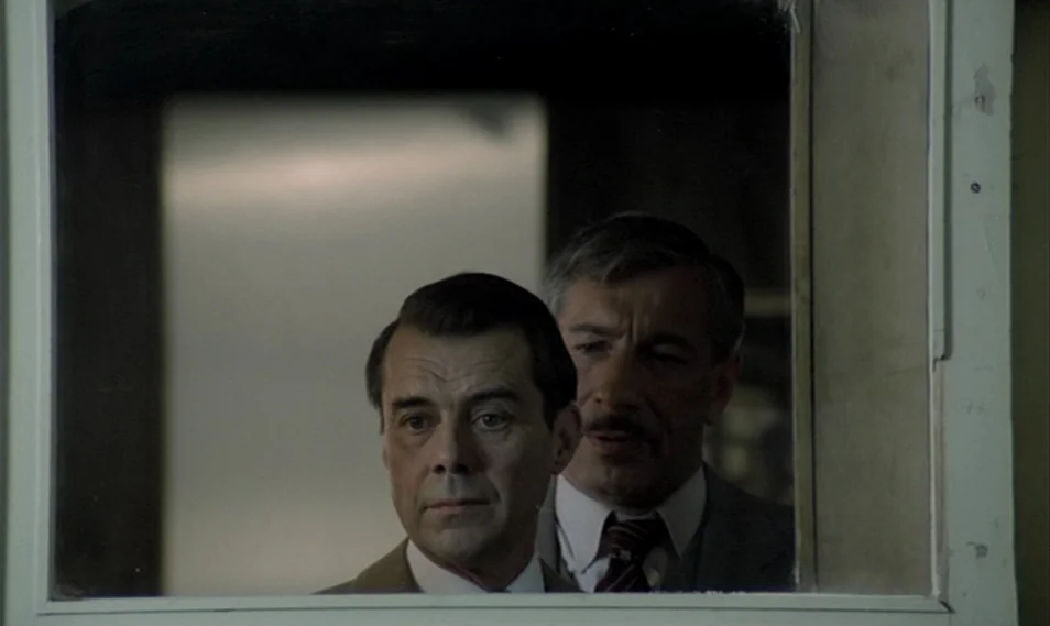 Despair (1978) movie still: closeup — Two middle-aged men stand together in a dimly lit, austere room with white walls. They observe their own…