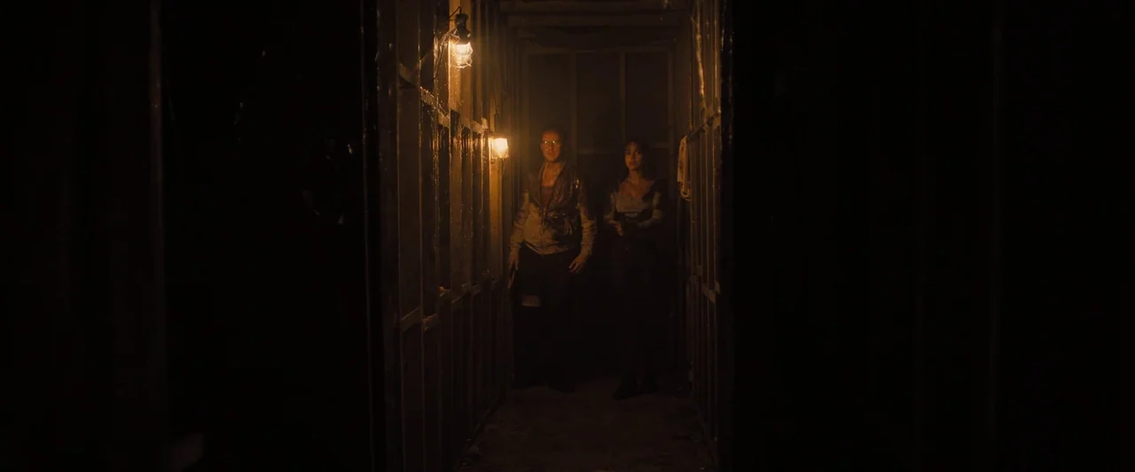 Abigail (2024) movie still: wide — A dimly lit narrow wooden passage with exposed stud wall construction. Two individuals stand in the shadows…