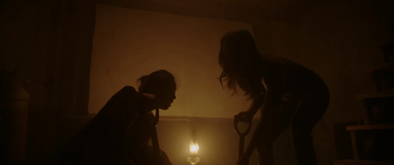 Impetigore (2019) movie still: medium — A dimly lit, windowless room with textured walls and a central oil lantern on the floor. Two figures in…