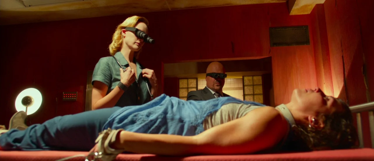 Rupture (2016) movie still: medium — A dimly lit room with deep red walls and industrial medical equipment present. A woman is restrained on a cot…