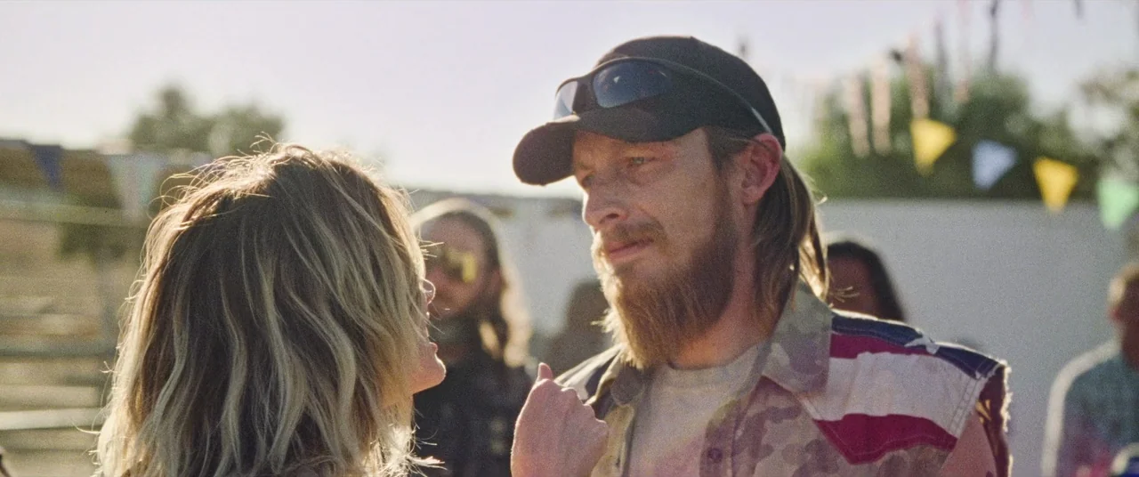 To Leslie (2022) movie still: medium — An outdoor fairground setting features blurred festive bunting in the background under bright daylight. A man…