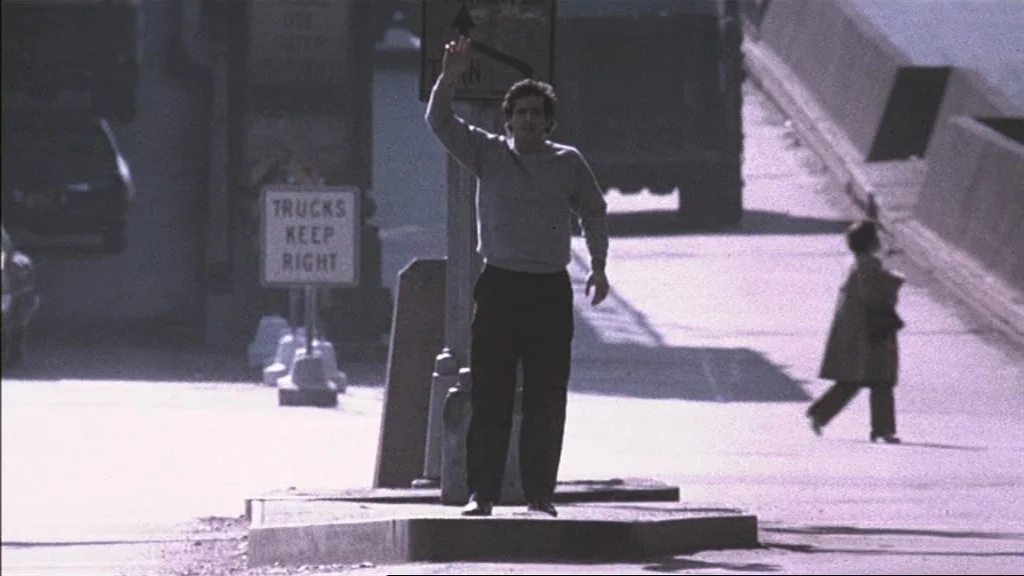 The Exorcist (1973) movie still: fullbody — A concrete traffic island sits in the middle of a paved city thoroughfare. A man in a grey sweatshirt…