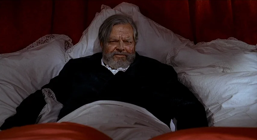 Malpertuis (1971) movie still: medium — A man lies against white pillows framed by heavy, deep red velvet curtains. The subject rests against the…