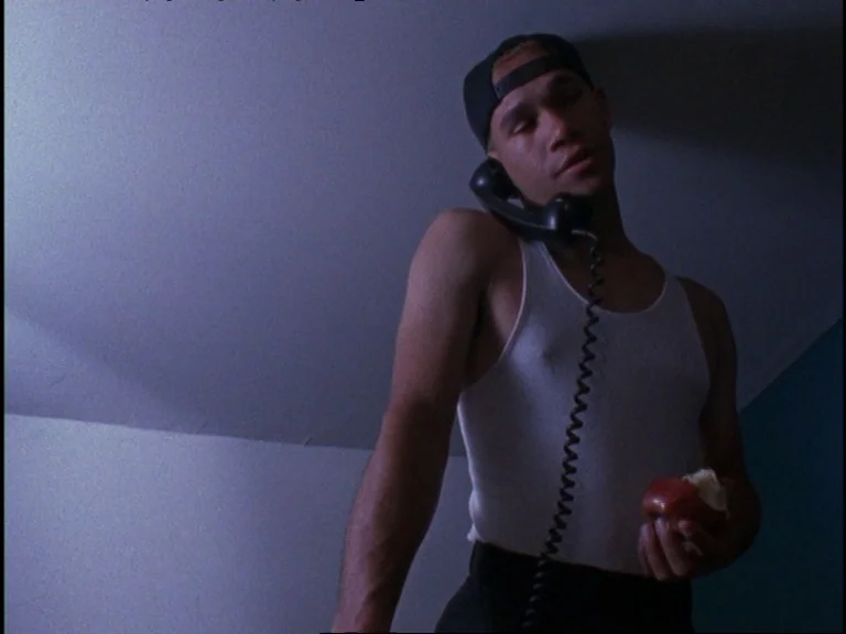 Totally F***ed Up (1993) movie still: medium — A dark corner of a room with muted blue and grey walls. A young man wearing a tank top talks on an…