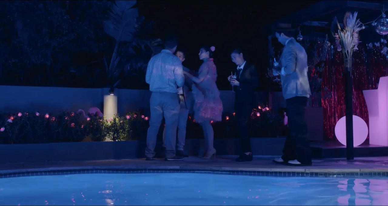 Five Blind Dates (2024) movie still: wide — A group of young adults stands around a blue lit pool at night. They stand in a casual cluster while…