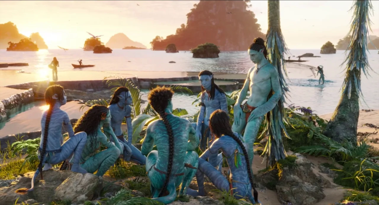 Avatar: The Way of Water (2022) movie still: wide — A group of blue-skinned humanoids sits on a tropical island beach at sunset. Rock formations…