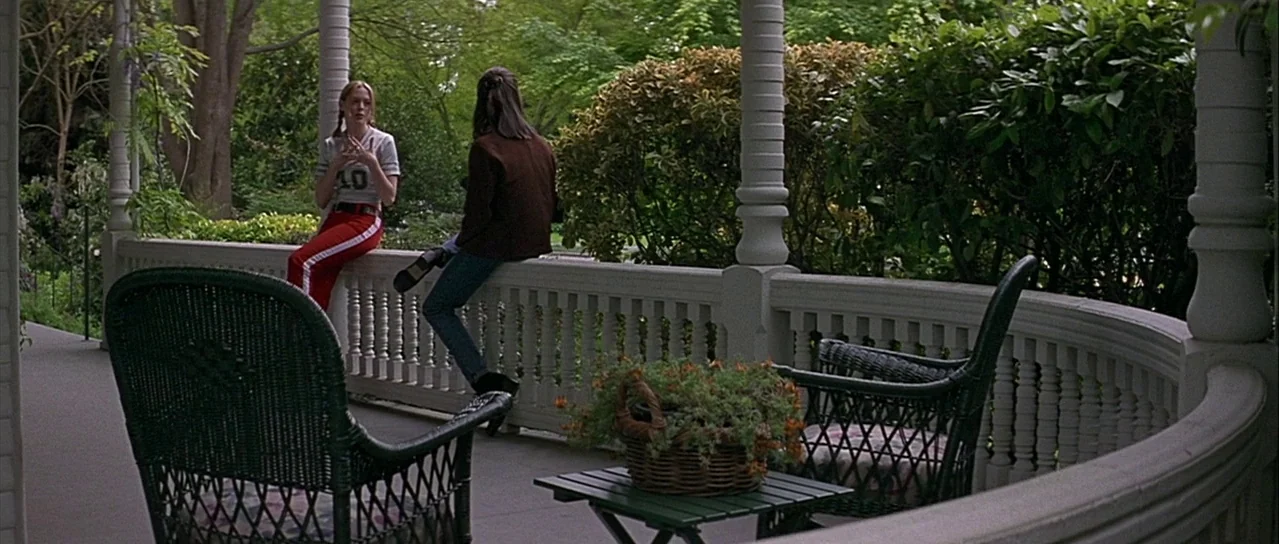 Scream (1996) movie still: wide — A wrap-around porch with white railings and wicker furniture overlooks lush green bushes. Two teenage girls sit on…