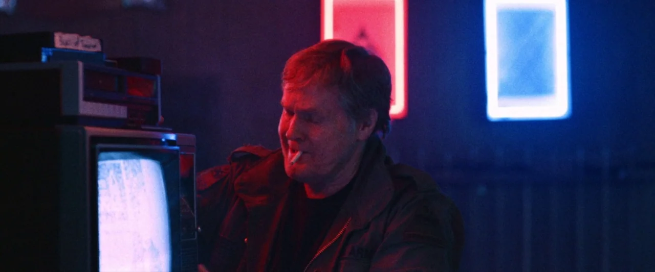 VFW movie still: medium — A dark space featuring stacked electronic equipment including a glowing television monitor. A man in a field jacket stands…