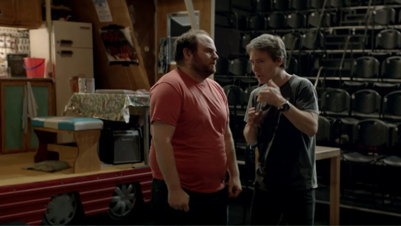 I Married a Dumbass (2016) movie still: medium — A cluttered rehearsal room features stacked chairs and a small wooden platform stage. Two men stand…