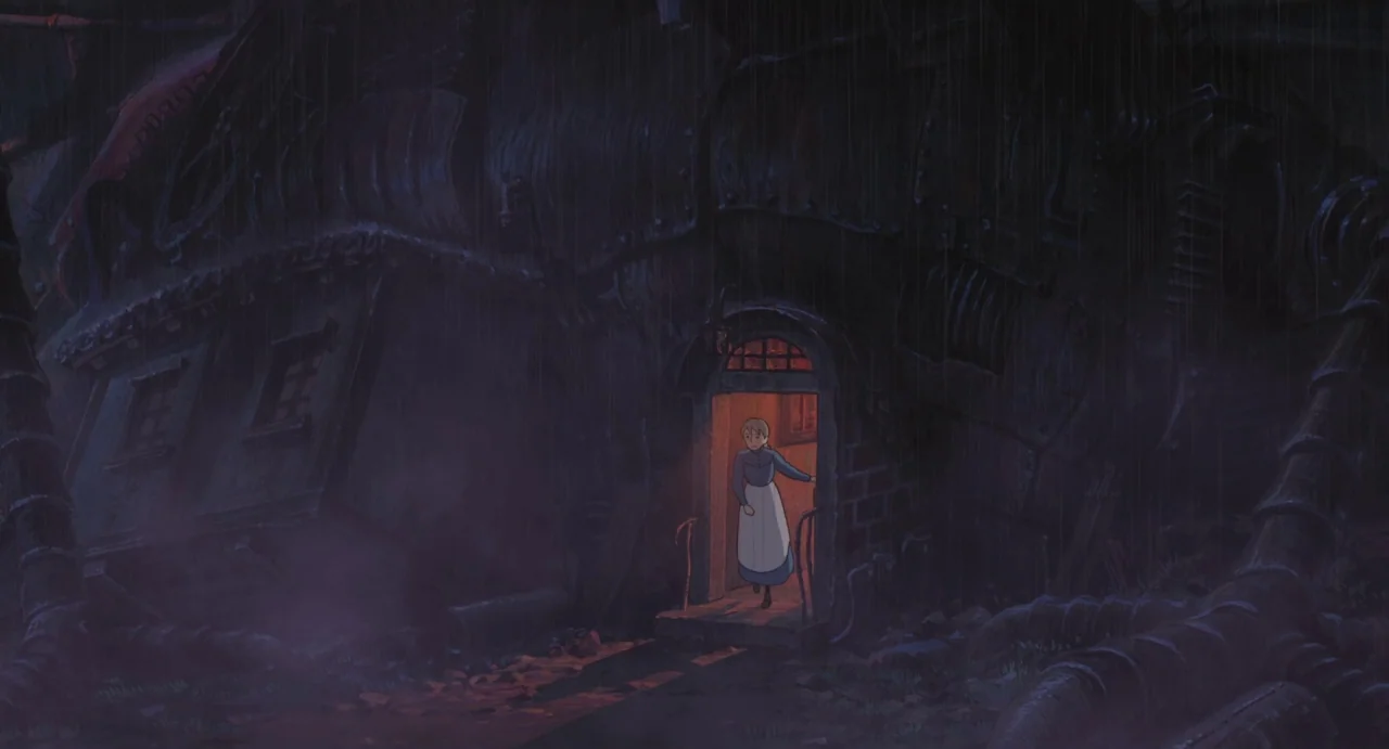 Howl’s Moving Castle (2004) movie still: wide — A dark, industrial structure made of heavy metal plates sits in the heavy rain. A young woman steps…