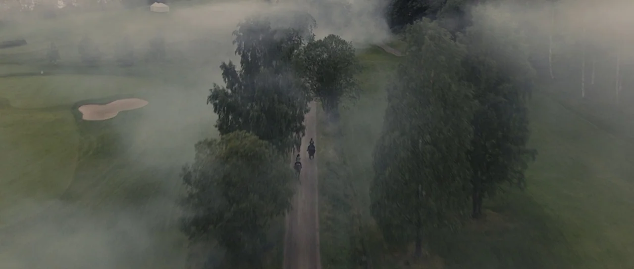 Melancholia (2011) movie still: wide — A golf course layout is blanketed in thick morning fog among trees. Two riders on horseback travel along a…