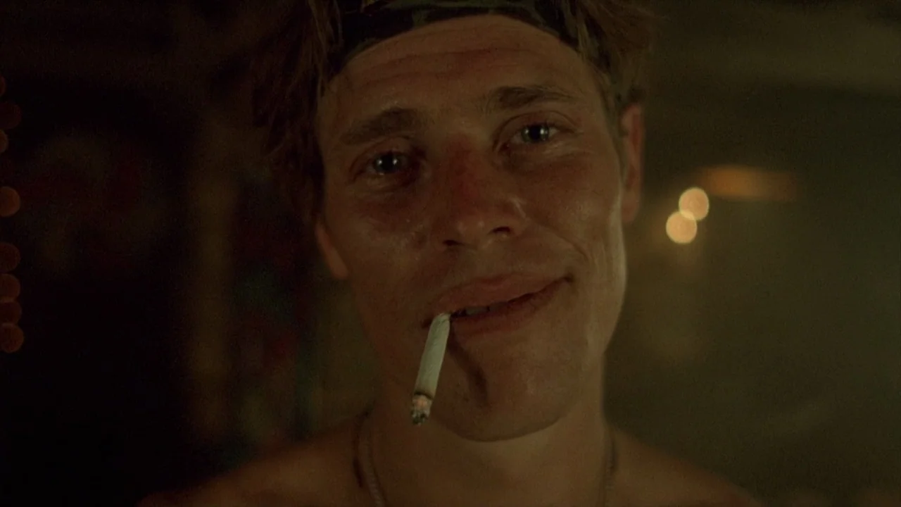 Platoon (1986) movie still: closeup — A dimly lit indoor space with indistinct dark surroundings. A young man stares into the lens with a cigarette…