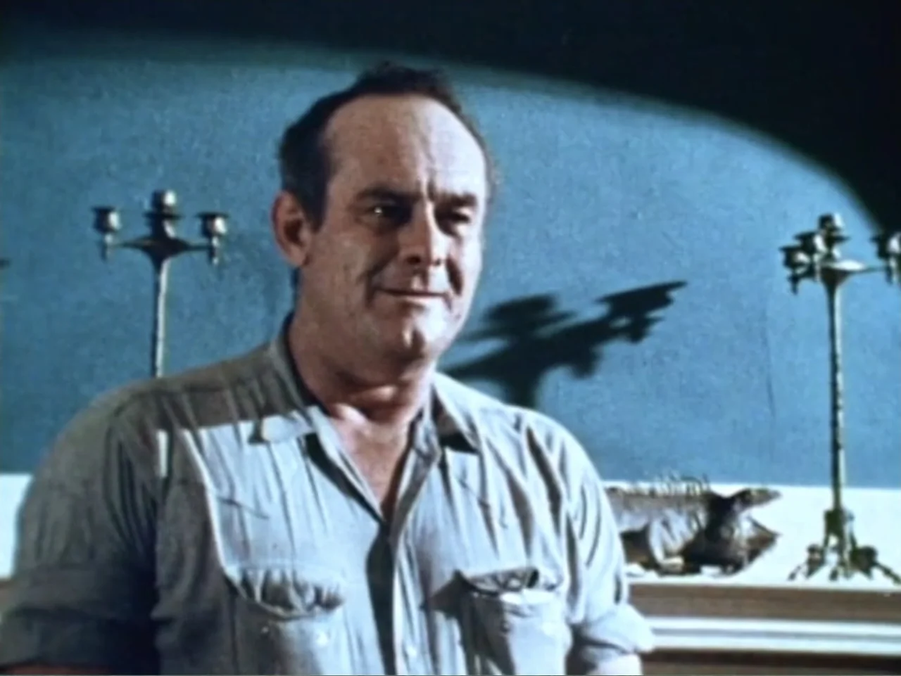 It's Alive (1974) movie still: medium — A dimly lit room features a blue wall adorned with two ornate candle holders. An adult man wearing a…