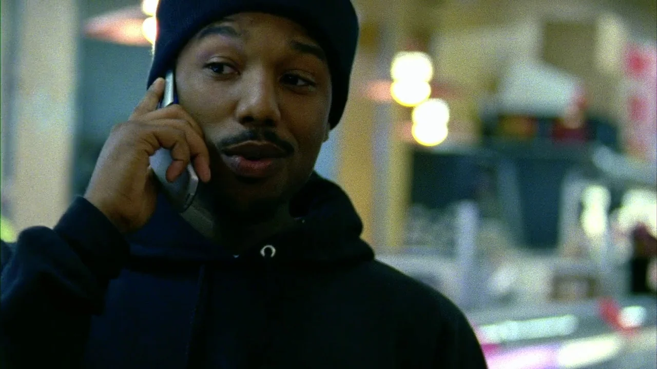Fruitvale Station (2013) movie still: closeup — A man in a dark hoodie stands inside a commercial interior space. The man holds a mobile phone to…