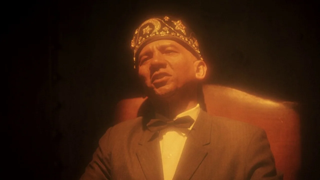 Malcolm X (1992) movie still: medium — A dimly lit, shadowed space with a dark background and a single leather chair. A man wearing a decorative fez…
