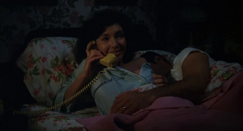 Melvin & Howard (1980) movie still: medium — A dark room features a bed with floral-patterned sheets and pillows. A woman lies in bed holding a…