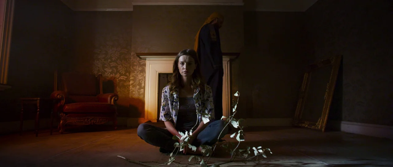 A Dark Song (2016) movie still: medium — A dimly lit room features patterned wallpaper and a dark leather armchair. A woman sits on the floor while…
