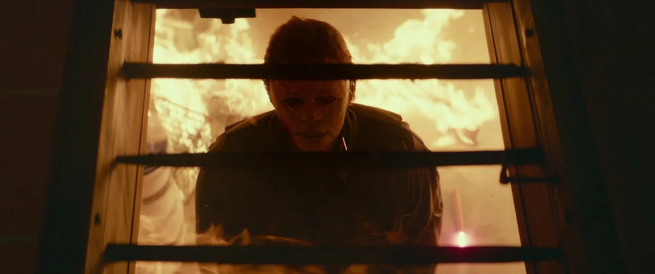 Halloween (2018) (2018) movie still: medium — A man wearing a white mask stands behind a metal ladder during a fire. Flames engulf the space around…