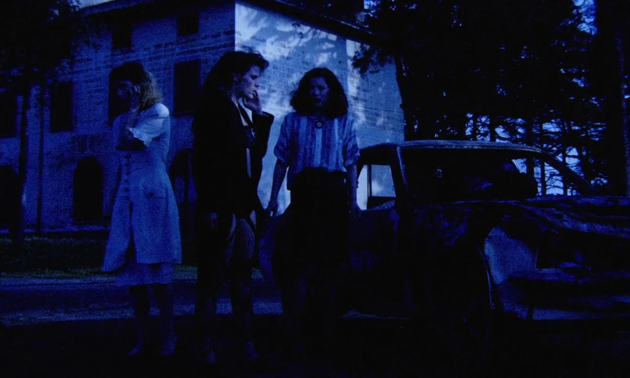 Paganini Horror (1989) movie still: fullbody — Three women stand together in the dark beside a damaged vehicle. A stone building with dark windows…