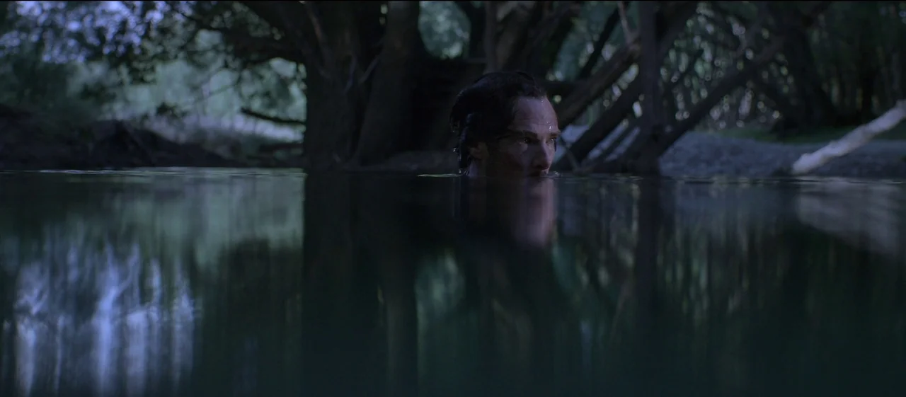 The Power of the Dog (2021) movie still: medium — A dark river flows through a dense wooded area during twilight hours. A man is submerged in the…