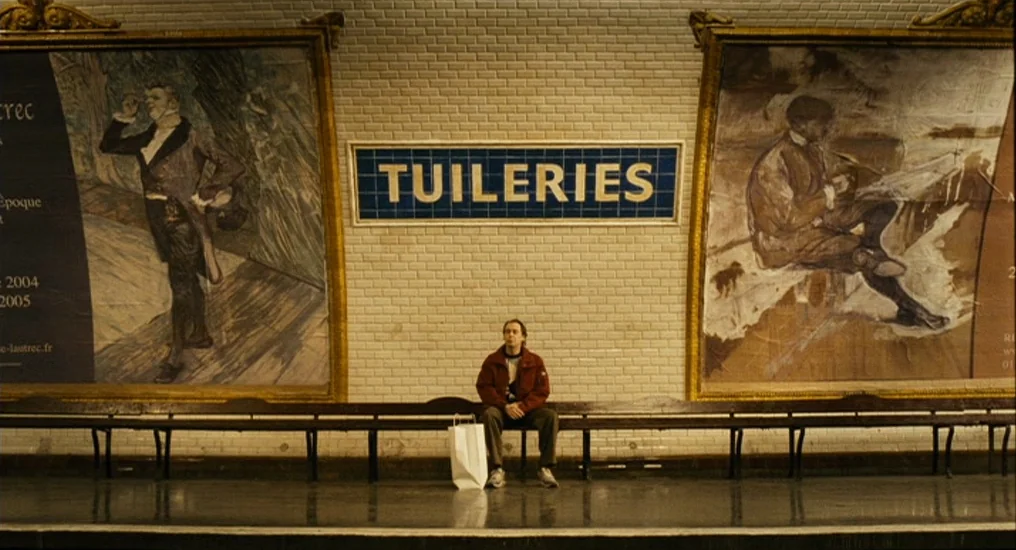 Paris, Je T’Aime (2006) movie still: wide — A Paris metro station platform features white tiled walls and large framed posters. A solitary man sits…